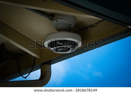 A modern security camera with a dark dome lens is attached to the underside of a building's eaves, monitoring the surroundings