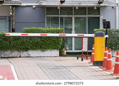 Modern security gate blocks entry to a building. Red and white barrier arm stands still, flanked by orange traffic cones. Green hedges line the sidewalk near glass windows