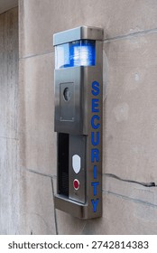 Modern security intercom panel mounted on a stone wall. Stainless steel emergency call box with surveillance camera, speaker, red call button and blue flashing light on top. The word “SECURITY” is displayed vertically on the side. Concept of safety, surveillance, access control and building protection