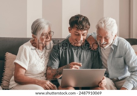 Modern senior couple learning computer technology from experienced young grandson, handsome smiling boy teaching to his grandparents how to use computer, make online shopping, book travel and vacation