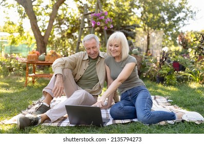 Modern Senior Spouses Shopping Online With Laptop, Sitting On Blanket In Garden, Enjoying Warm Spring Day. Elderly Couple Surfing Internet Or Chatting With Family