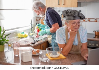 Modern Senior Woman With Chef Hat Preparing An Home Made Cake Looking On Web For The Recipe While Her Husband Washes The Dishes. Concept Of Family And Domestic Partnership