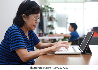 Modern senior woman using a portable digital tablet at home, grandchild using a computer as background. Family E-learning concept.