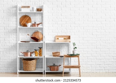 Modern shelf units with kitchenware and stand near white brick wall