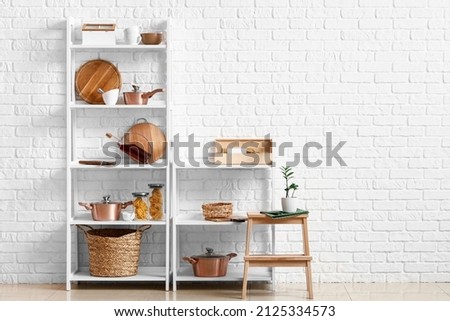 Modern shelf units with kitchenware and stand near white brick wall