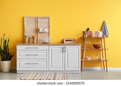 Modern shelving unit with dishware, kitchen counter and houseplant near yellow wall