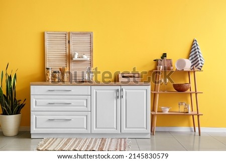 Modern shelving unit with dishware, kitchen counter and houseplant near yellow wall