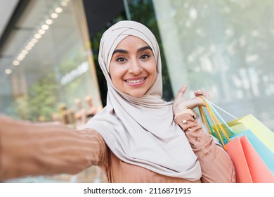 Modern shopping with gadget, blogger and buying clothes in city, sale. Smiling young arab lady in hijab with colored bags with purchases looking at webcam and taking photo near shop, copy space