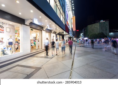 Modern Shopping Street And People In Urban City With Cement Road At Night  