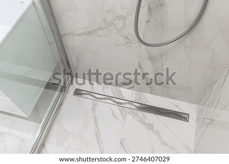 A modern shower area with marble-patterned tiles, linear stainless steel drain, glass door, and flexible chrome shower hose mounted on the wall.