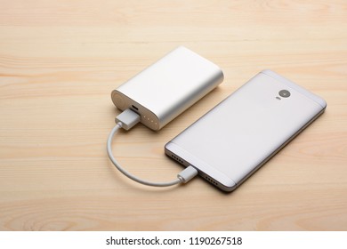 Modern Silver Smartphone Lays On Light Wooden Table With Its Back Panel Up While Charging With Power Bank