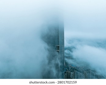 Modern skyscraper tower emerging from dense clouds above a cityscape. Aerial view of urban architecture shrouded in fog and mist with river visible below.