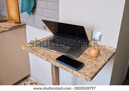 Modern small kitchen setup featuring granite countertop with open laptop, smartphone, and red apple. Soft natural lighting enhances neutral colored environment