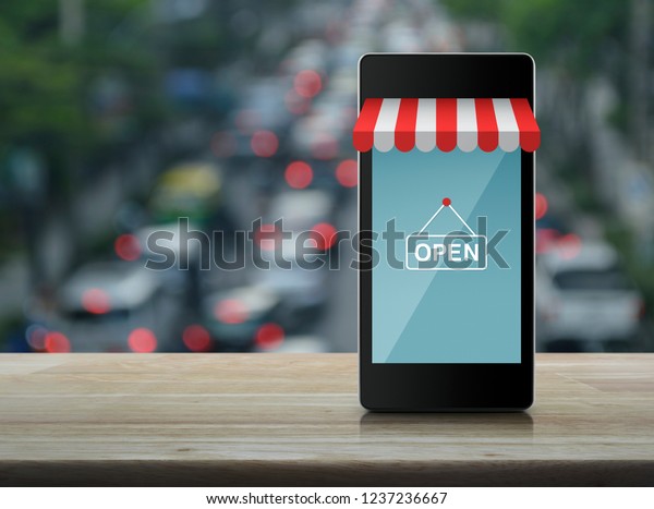 Modern smart mobile phone with online\
shopping store graphic and open sign on wooden table over blur of\
rush hour with cars and road, Business shop online\
concept