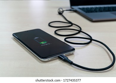 Modern smartphone are charging from the laptop on a light wooden table. Modern technology concept. Selective focus