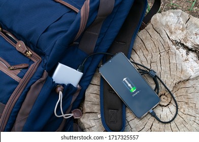 Modern Smartphone Are Charging From Power Bank Lies Next To The Backpack On A Wooden Stump. Modern Technology Concept