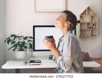 Modern Smiling Beautiful Woman Working And Drinking Coffee From Reusable Mug At Home Office. Minimalist Slow Life Style Workplace Of Creative Female In Glasses And Earphones Using Computer Technology.