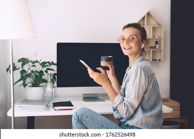 Modern smiling beautiful woman working and drinking coffee from reusable mug at home office. Minimalist slow life style workplace of creative female in glasses and earphones using computer technology.