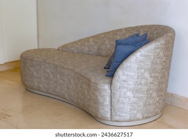 Modern sofa with a unique curved shape is placed against a white wall, and the floor is tiled. It is upholstered in a light colored fabric that has a distinct texture. A blue cushion rests on the sofa