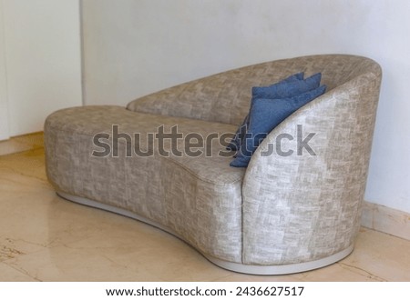 Modern sofa with a unique curved shape is placed against a white wall, and the floor is tiled. It is upholstered in a light colored fabric that has a distinct texture. A blue cushion rests on the sofa