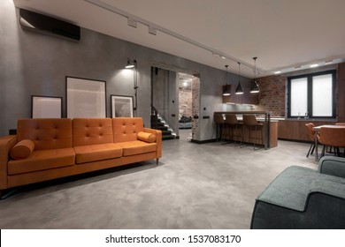 Modern Spacious Interior Of Studio Flat Including Brown Wooden Kitchen With Dinning Furniture Sets And Soft Orange Couch With Gray Armchair Against Red Brick And Gray Plastered Walls In Loft Style