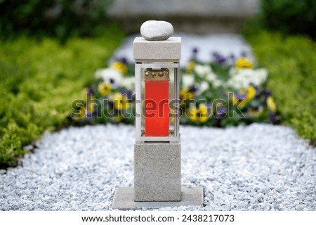 modern square grave lantern with red candle on white gravel  in front of plants in blurred background