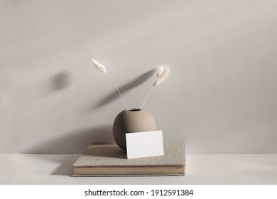 Modern stationery still life scene. Beige spheric vase with dry lagurus grass. Table background in sunlight. Blank business card mockups lean on old book. Champagne wall, long shadows. 