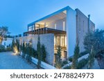 A modern stone villa with a white facade, lit up at dusk. The villa features a wooden pergola, creating a shaded patio area. A stone wall surrounds the property, with potted plants lining the walkway