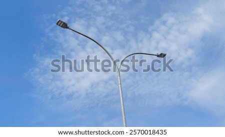 Modern street light with two rectangular LED arms against a bright blue sky with scattered clouds. Tall, slender pole in a clean, minimalist outdoor scene, highlighting urban design simplicity