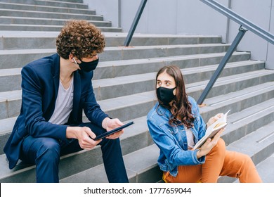 Modern students. Distance learning. Social distance. New normal life. Young hipsters girl and boy students in facial masks, reads book while sitting on stairs outdoor with a tablet computer. - Powered by Shutterstock