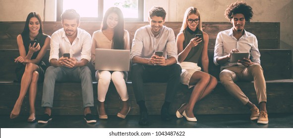 Modern Students. Front View Of Young People Using Different Gadgets While Sitting Close To Each Other In A Row