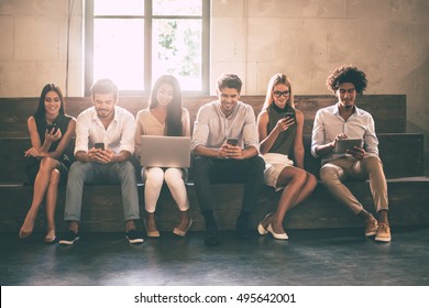 Modern Students. Front View Of Young People Using Different Gadgets While Sitting Close To Each Other In A Row 