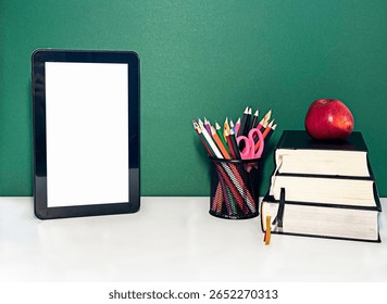 A modern study setup featuring a tablet, a cup of colorful pens, an apple, and stacked books on a white desk against a green wall. High quality photo