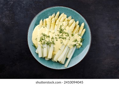 Modern style traditional steamed white asparagus with sauce hollandaise served herbs as top view on a Nordic design plate on a rustic black board 