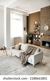 Modern Stylish Living Room With Large Windows And Sofa With Pillow And Plaid On The Backgroung Of Brown Wall With Brick Fireplace, Shelving With Books, Decor And Potted Plants. Cozy Chalet Interior.