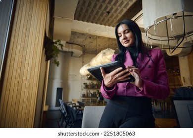 Modern stylish middle-aged successful woman entrepreneur, sales manager, chief executive working remotely on a digital tablet from the interior of a restaurant. People and business. Low angle view - Powered by Shutterstock