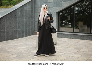 Modern Stylish Muslim Woman In Hijab, Leather Jacket And Black Abaya Walking In City Street , Wearing Sunglasses And Bag, Fashion Style Trend