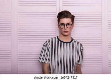 Modern stylish young man hipster in fashionable glasses with a hairstyle in a trendy striped t-shirt posing near a vintage wooden pink wall in the studio. Portrait of a attractive guy. English fashion