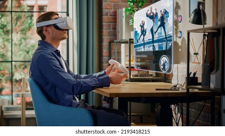 Modern Successful Man Sitting In Living Room At Home, Using Virtual Reality Headset With Controllers To Check Social Media Application. Young Male Watching A Dance Music Video Clip.