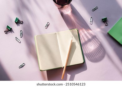 Modern summer stationery still life. A glass of water with long shadow and open notepad with pencil on purple background. Top view, flat lay.