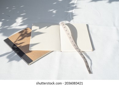 Modern summer stationery still life.Empty and open diary or notepad mockup with fabric bookmark with small embroidered hearts. Long shadows of sunset on the blank pages and on the white  tablecloth