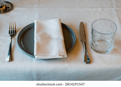 Modern table setting with ceramic plate folded white napkin knife fork and drinking glass viewed from above.