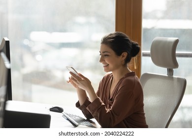 Modern Tech In Office. Happy Young Mixed Race Woman Manager Sit At Desk By Computer Messaging On Cell Distracted From Work On Pc. Smiling Indian Lady Corporate Employee Answer Phone Call At Workplace