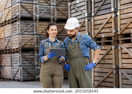 Modern tech for work in warehouse, motion control and logistic. Busy millennial lady in uniform and gloves shows tablet to guy worker in helmet in storage with many wooden containers, free space