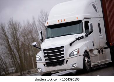 Modern Technically Equipped With Innovations White Big Rig Long Haul Semi Truck Tractor Transporting Container With Commercial Cargo On Flat Bed Trailer On Wet Dangerous Road In Rainy Storm Weather