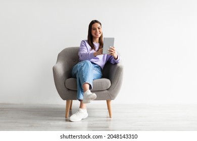 Modern technologies and communications. Full length of cheery young woman with tablet pc sitting in armchair, working or studying online, having video chat against white studio wall - Shutterstock ID 2048143310