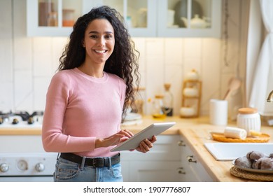 Modern Technologies In Daily Life. Portraif Of Happy Young Woman Standing With Digital Tablet In Kitchen Interior, Smiling At Camera, Positive Lady Browsing Social Media While Relaxing At Home