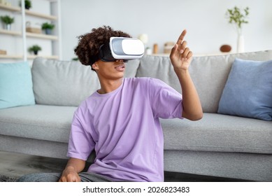 Modern Technologies And Entertainments. Cool Black Teen Guy In VR Headset Exploring Virtual Reality At Home, Touching Imaginary Screen, Interacting With Cyberspace