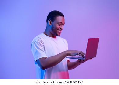 Modern Technologies. Happy Black Man Using Laptop Computer In Neon Light, Enjoying Online Communication, Gaming And Remote Job Offers, Excited Guy Standing Over Purple Studio Background, Copy Space