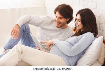 Modern technologies and pregnancy. Happy pregnant woman and her husband using digital tablet while lying on bed at home